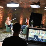 Eine Person in der Regie, die auf ein Videopodcast-Setup blickt, in welchem zwei Personen sitzen.