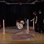 Eine Bühne mit Schlagzeug, Lichtröhren und Teppichen drauf, auf welcher zwei Produzentinnen das Setup besprechen.