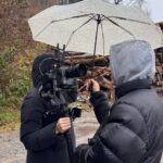 Eine Person mit Kapuze, die schützend einen Regenschirm über die Kamerafrau hält.