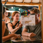 Eine Person mit Filmklappe zwischen den Schauspielenden, welche die nächste Szene ansagt.