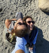 Zwei Freudinnen, die sich am Strand in der Sonne gegenseitig aneinander anlehnen und lachen.