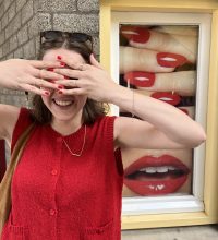 Eine Person verdeckt das Gesicht mit den Händen und zeigt die rotlackierten Fingernägel, die ähnlich aussehen wie auf dem Plakat dahinter.