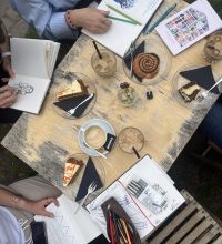 Tisch mit fünf Leuten voller Kaffee und Kuchen und Sketchbuechern.