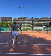 Eine Person in Sportkleidung mit Tennisschläger in der Hand auf dem Weg auf den Tennisplatz.